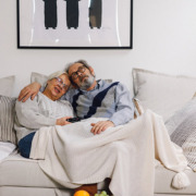 An older couple cuddled together under a blanket on the couch.