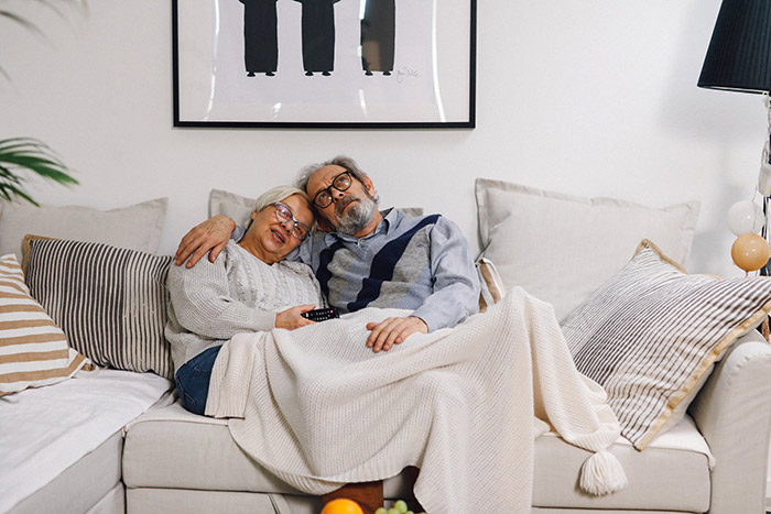 An older couple cuddled together under a blanket on the couch.